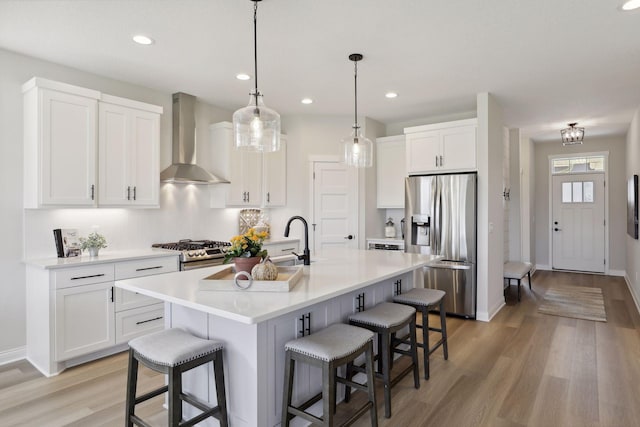 kitchen featuring white cabinets, light countertops, wall chimney range hood, appliances with stainless steel finishes, and an island with sink