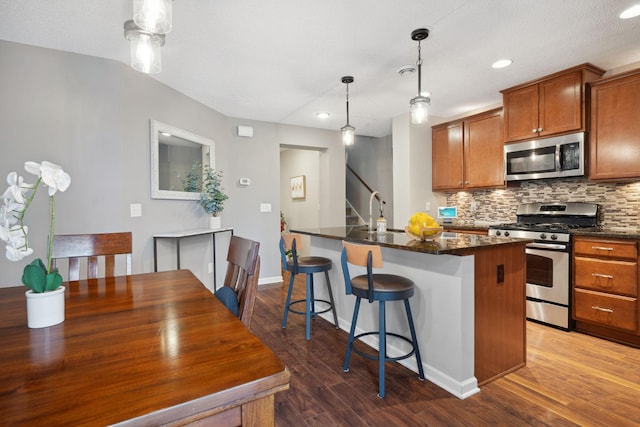 kitchen with brown cabinetry, wood finished floors, a center island with sink, stainless steel appliances, and decorative backsplash