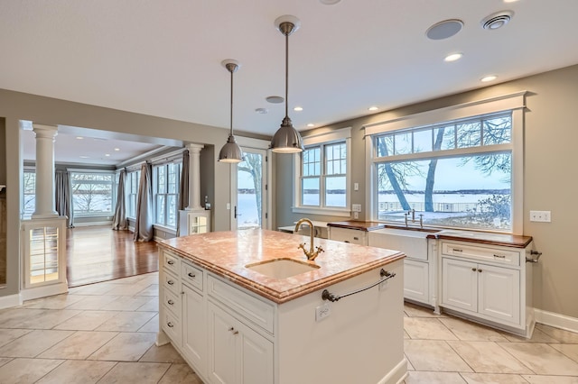 kitchen with sink, ornate columns, an island with sink, pendant lighting, and white cabinets