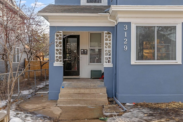 property entrance with stucco siding, fence, and roof with shingles