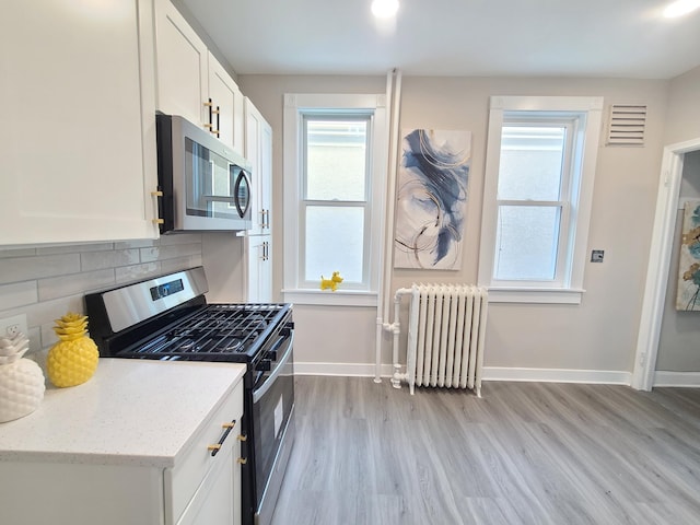 kitchen with appliances with stainless steel finishes, radiator heating unit, white cabinetry, decorative backsplash, and light stone countertops