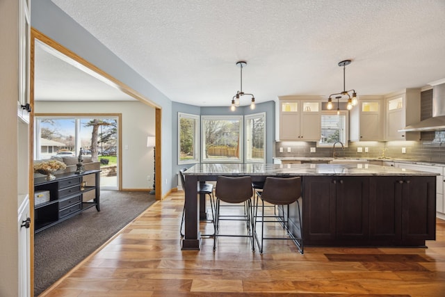kitchen with wall chimney range hood, tasteful backsplash, a center island, light wood-style floors, and glass insert cabinets