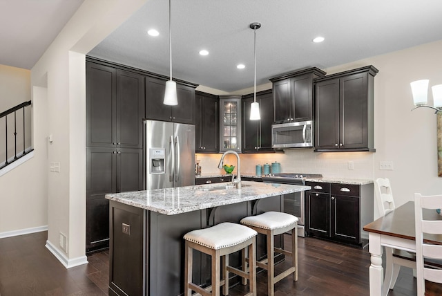 kitchen with pendant lighting, appliances with stainless steel finishes, a kitchen island with sink, light stone counters, and tasteful backsplash