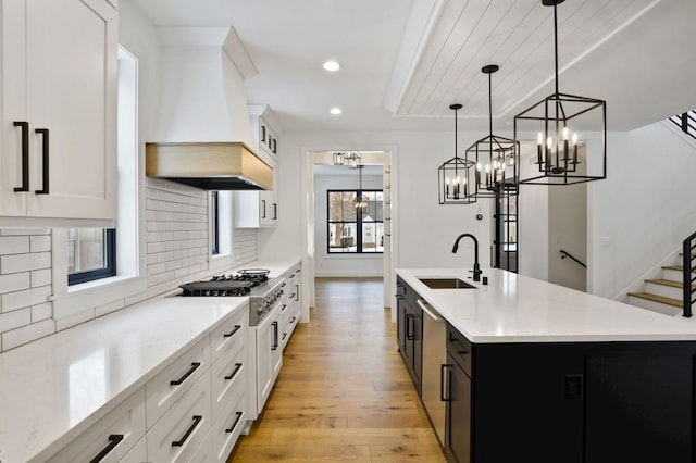 kitchen featuring white cabinets, custom range hood, decorative light fixtures, a kitchen island with sink, and a sink