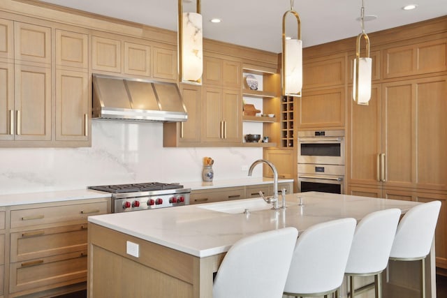 kitchen with sink, light stone counters, an island with sink, stainless steel double oven, and wall chimney range hood