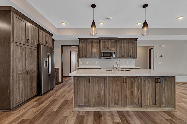 kitchen with stainless steel appliances, dark brown cabinetry, light stone countertops, a center island with sink, and decorative light fixtures