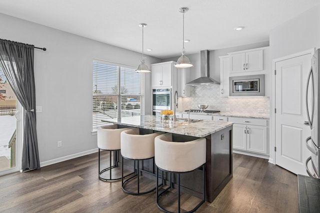 kitchen with an island with sink, appliances with stainless steel finishes, white cabinets, and wall chimney range hood