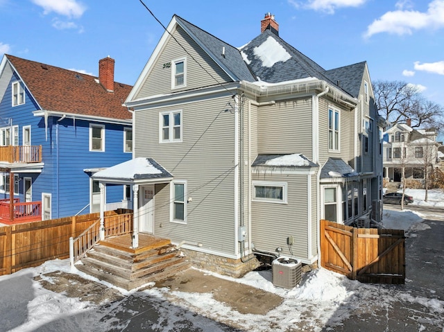 snow covered house with central AC, a shingled roof, a chimney, and fence