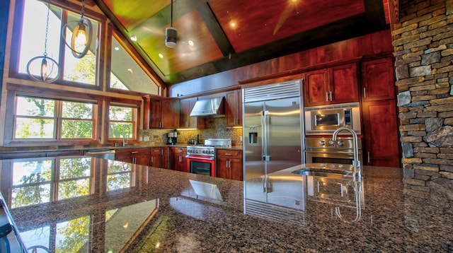 kitchen featuring reddish brown cabinets, dark stone countertops, built in appliances, hanging light fixtures, and wall chimney range hood