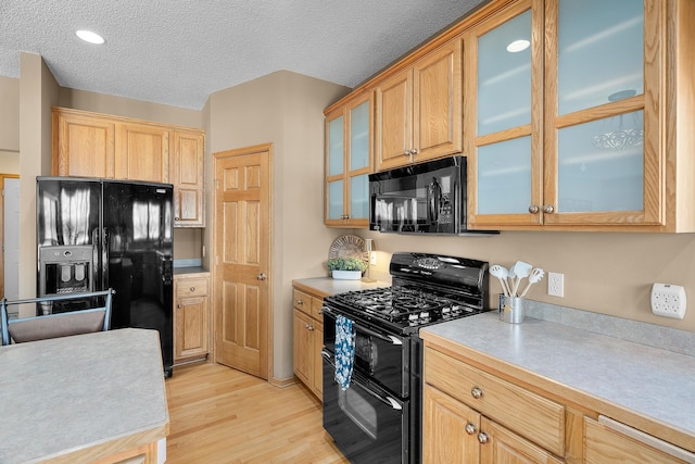 kitchen featuring a textured ceiling, light countertops, black appliances, light wood finished floors, and glass insert cabinets