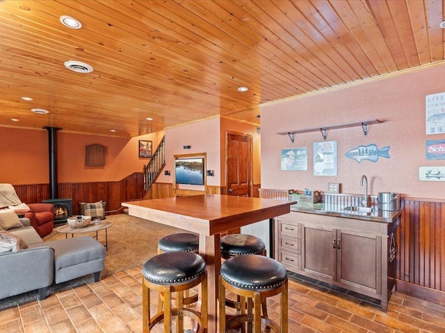 kitchen with wainscoting, wooden ceiling, a wood stove, a sink, and ornamental molding