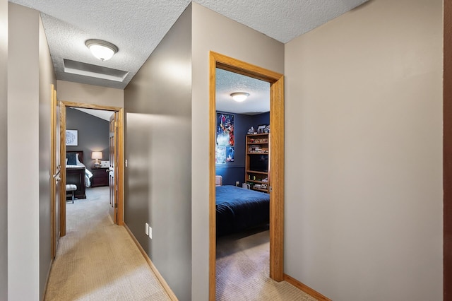 hall featuring attic access, light colored carpet, a textured ceiling, and baseboards