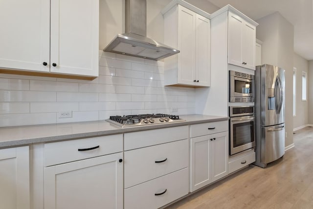 kitchen featuring wall chimney range hood, light hardwood / wood-style flooring, appliances with stainless steel finishes, white cabinetry, and decorative backsplash