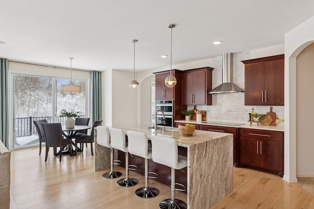 kitchen with arched walkways, tasteful backsplash, double oven, light wood-style floors, and wall chimney exhaust hood