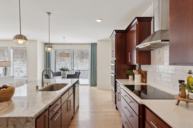 kitchen with a sink, wall chimney range hood, appliances with stainless steel finishes, light wood-type flooring, and tasteful backsplash