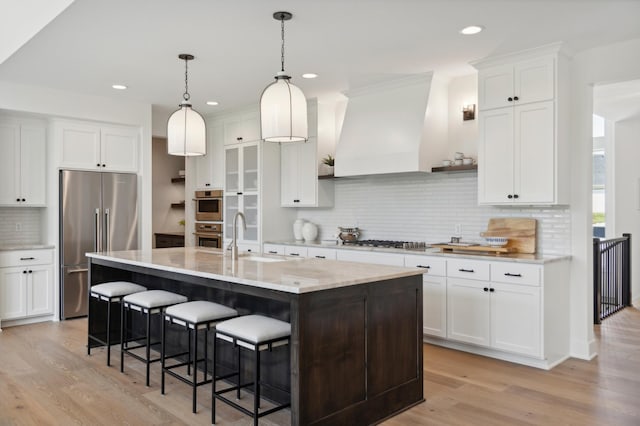 kitchen featuring sink, custom exhaust hood, white cabinetry, an island with sink, and stainless steel appliances