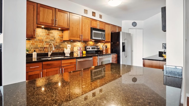 kitchen featuring a sink, appliances with stainless steel finishes, backsplash, brown cabinets, and dark stone counters