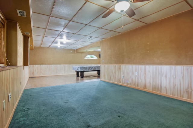 interior space with a drop ceiling, wood walls, ceiling fan, and carpet