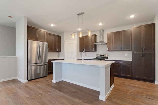 kitchen with sink, appliances with stainless steel finishes, an island with sink, decorative light fixtures, and wall chimney exhaust hood