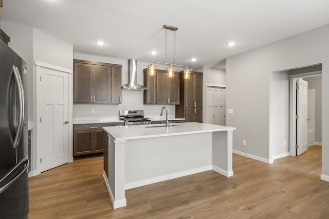 kitchen with sink, hanging light fixtures, appliances with stainless steel finishes, a kitchen island with sink, and wall chimney range hood