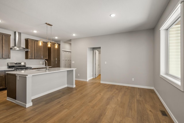 kitchen with sink, decorative light fixtures, an island with sink, stainless steel stove, and wall chimney range hood