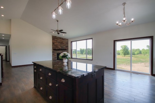 kitchen with pendant lighting, dark wood-type flooring, ceiling fan with notable chandelier, dark stone countertops, and a stone fireplace