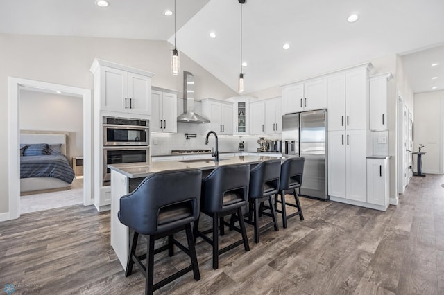 kitchen featuring a kitchen island with sink, a sink, appliances with stainless steel finishes, wall chimney exhaust hood, and glass insert cabinets