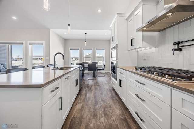 kitchen featuring stainless steel gas cooktop, a center island with sink, hanging light fixtures, white cabinetry, and wall chimney exhaust hood