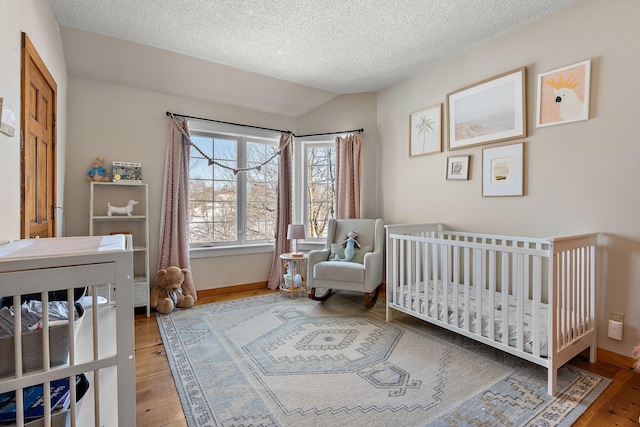 bedroom with vaulted ceiling, a textured ceiling, wood finished floors, a nursery area, and baseboards