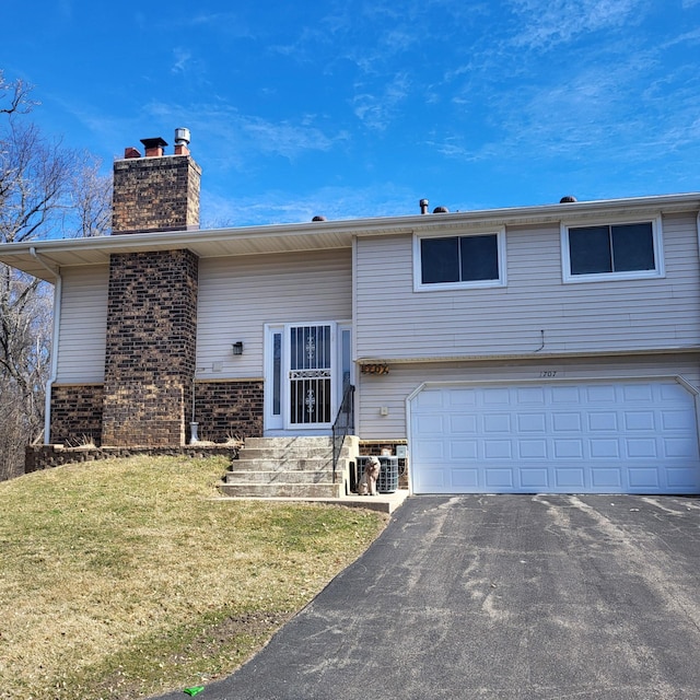 bi-level home featuring aphalt driveway, brick siding, a chimney, a garage, and a front lawn