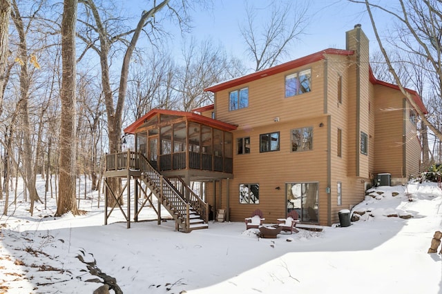 snow covered house with stairs, a chimney, a wooden deck, and a sunroom