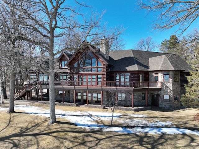 snow covered rear of property with a shingled roof, log siding, a chimney, a deck, and stone siding