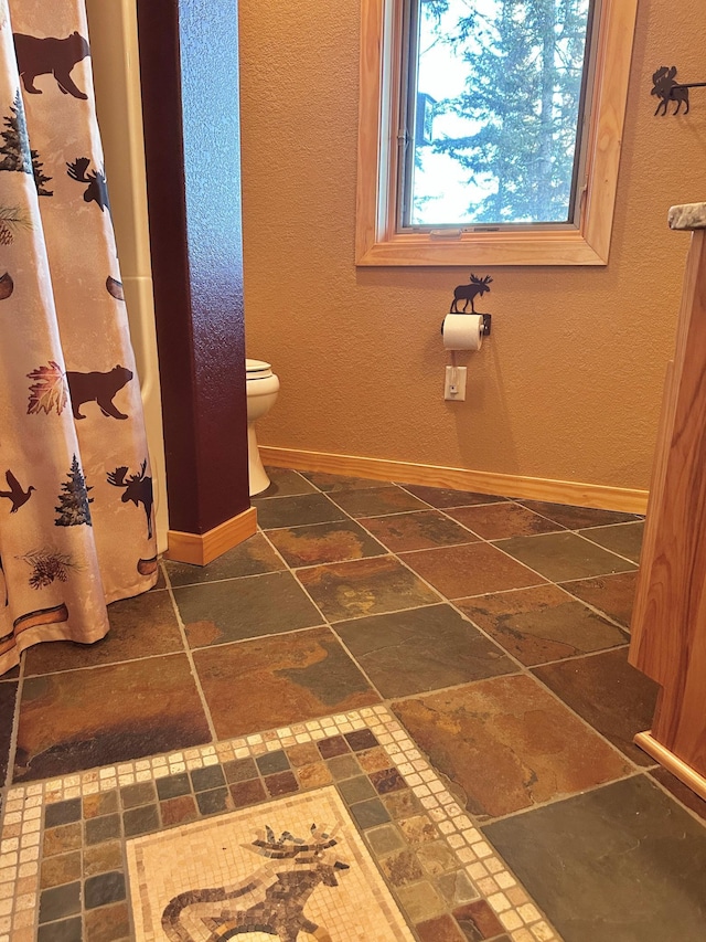 bathroom with curtained shower, baseboards, toilet, and stone tile floors