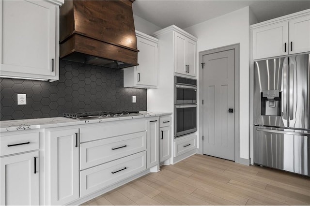 kitchen featuring white cabinets, stainless steel appliances, light stone countertops, custom range hood, and light wood-type flooring