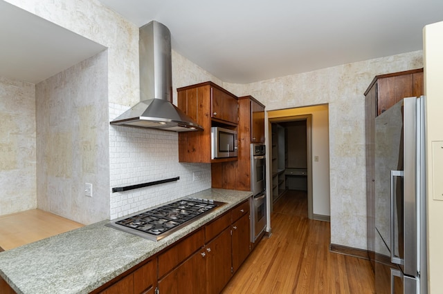 kitchen with wall chimney exhaust hood, appliances with stainless steel finishes, light countertops, light wood-style floors, and backsplash