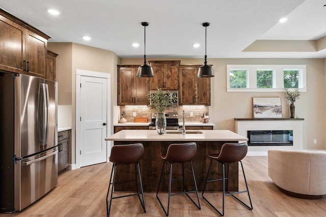 kitchen with light wood-type flooring, a kitchen island with sink, stainless steel appliances, hanging light fixtures, and tasteful backsplash