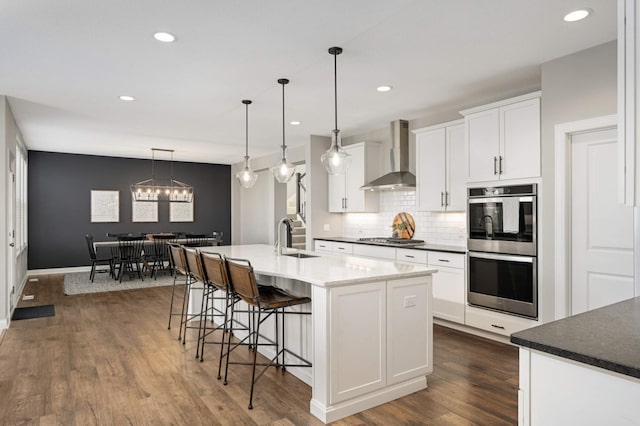 kitchen with sink, white cabinetry, an island with sink, pendant lighting, and wall chimney range hood