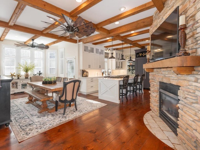 dining room with dark wood-style floors, a fireplace, a ceiling fan, and beamed ceiling