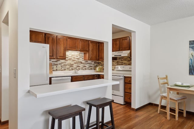 kitchen with under cabinet range hood, a peninsula, white appliances, wood finished floors, and a kitchen bar