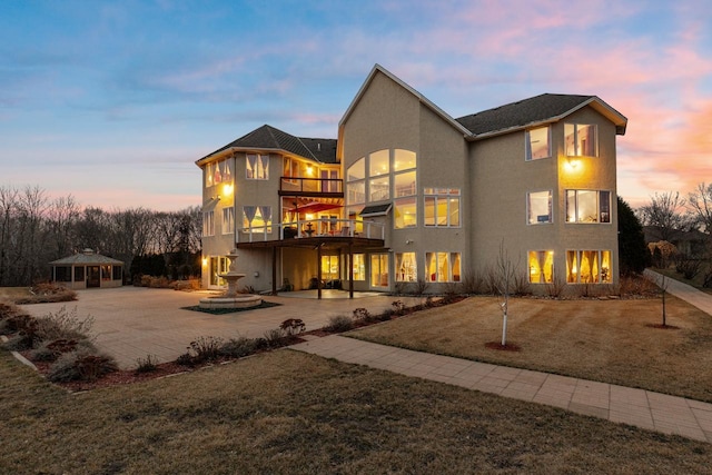 back of property at dusk featuring curved driveway, stucco siding, a patio, a lawn, and a gazebo