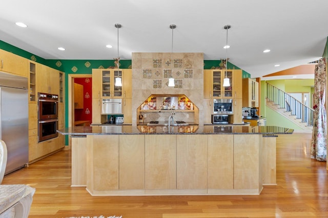 kitchen with dark stone counters, a large island with sink, glass insert cabinets, stainless steel appliances, and light wood finished floors