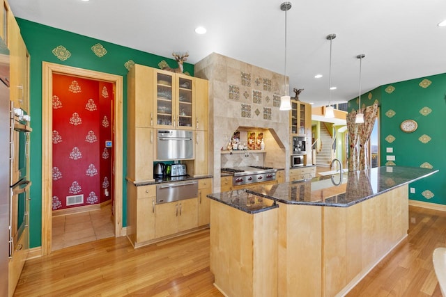 kitchen with light wood finished floors, a kitchen island with sink, light brown cabinets, and a warming drawer