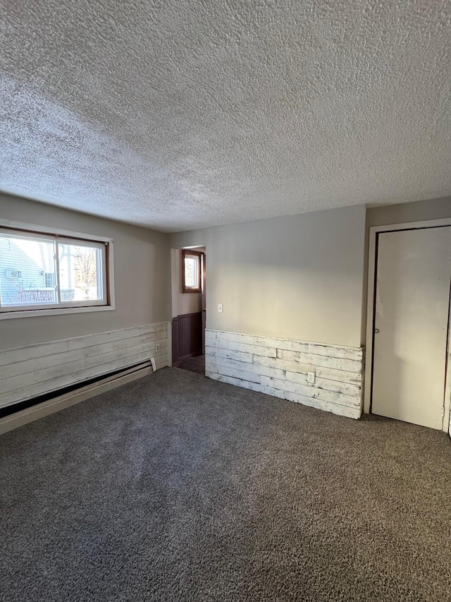 empty room featuring a textured ceiling, wooden walls, a wainscoted wall, carpet floors, and baseboard heating