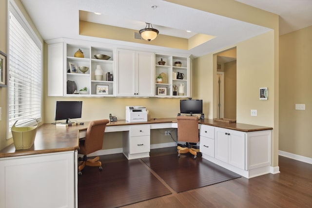 home office featuring visible vents, baseboards, built in study area, a tray ceiling, and dark wood finished floors