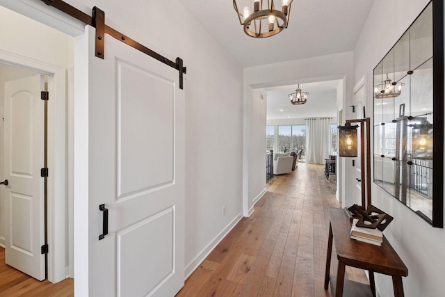 hallway featuring baseboards, a barn door, light wood-style flooring, and an inviting chandelier