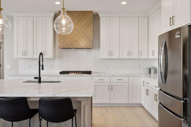 kitchen with sink, stainless steel refrigerator, white cabinetry, hanging light fixtures, and light stone counters