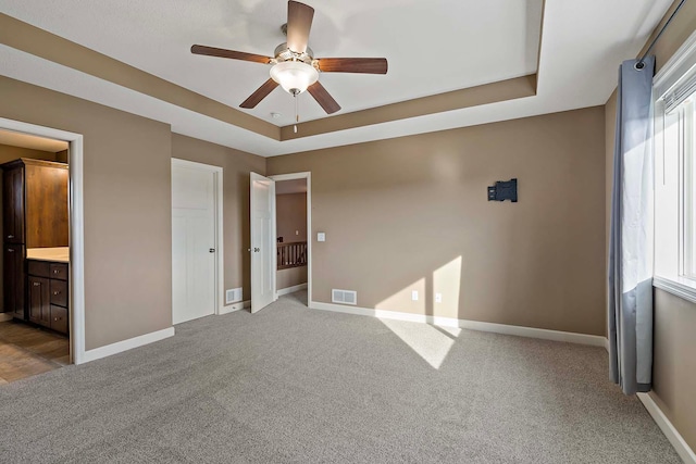 unfurnished bedroom featuring a tray ceiling, ensuite bath, ceiling fan, and carpet flooring