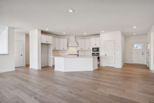 kitchen featuring built in microwave, custom range hood, stainless steel oven, light wood-type flooring, and backsplash