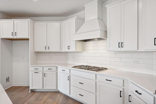 kitchen featuring custom exhaust hood, white cabinets, stainless steel gas stovetop, light wood-style floors, and tasteful backsplash