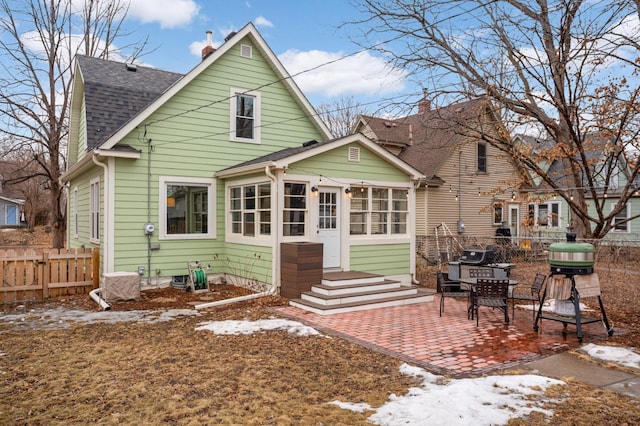 snow covered house featuring a shingled roof, a patio area, and fence